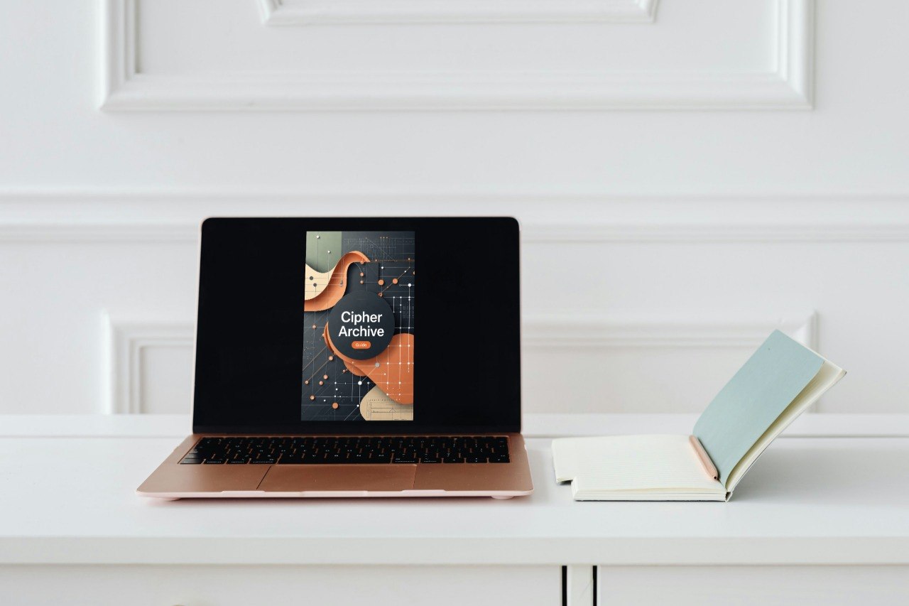 A laptop on a white desk displaying the book cover of 'Ciber Archive', with an open notebook beside it against a white paneled wall.