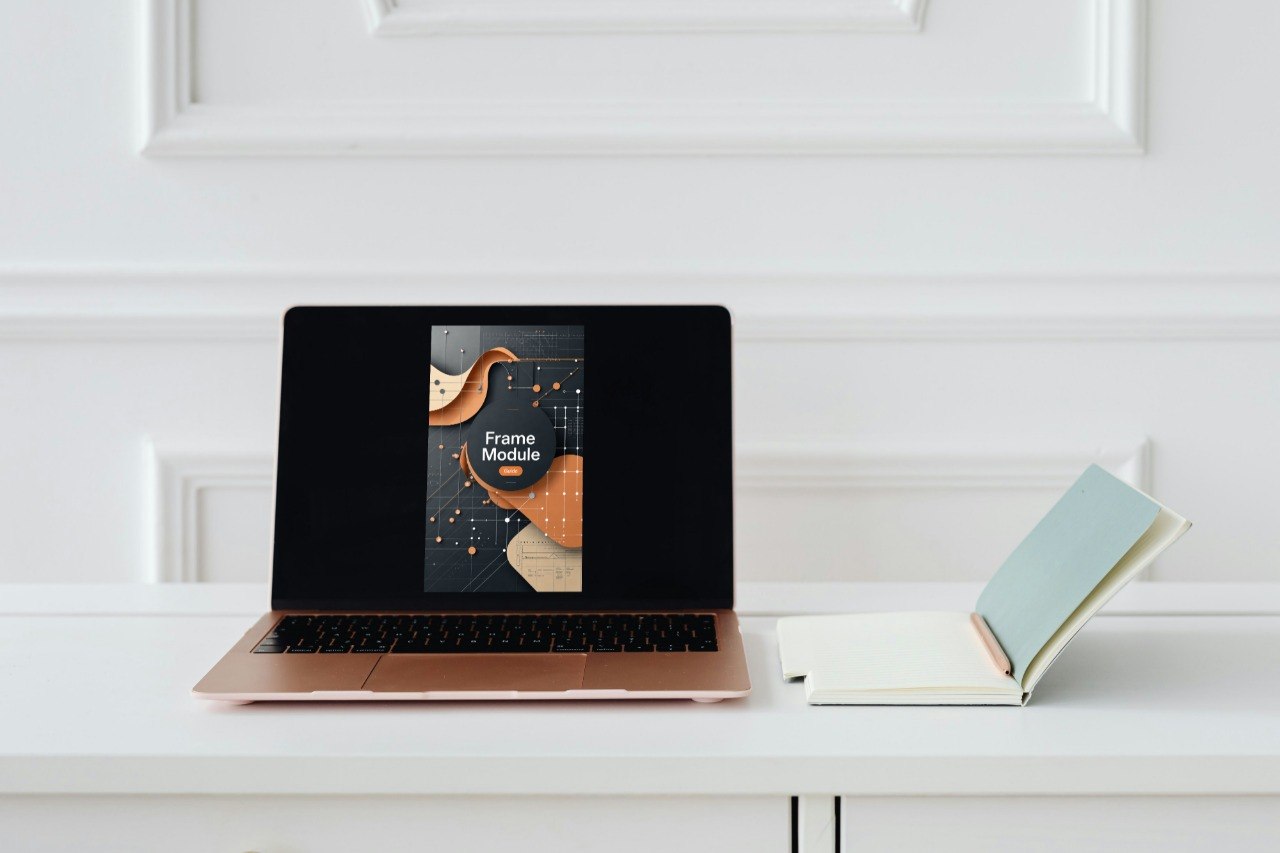 A laptop sits open on a white table against a white paneled wall, displaying a book cover mockup titled 'Frame Module.' An open notebook with a light blue cover rests beside it.